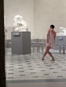 a ballerina in mid step in a marble hall art gallery with marble rodin statues