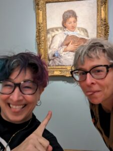 liz and sister grinning, sticking out tongues cheerfully while liz points at famous Cassat painting of a lady with a book