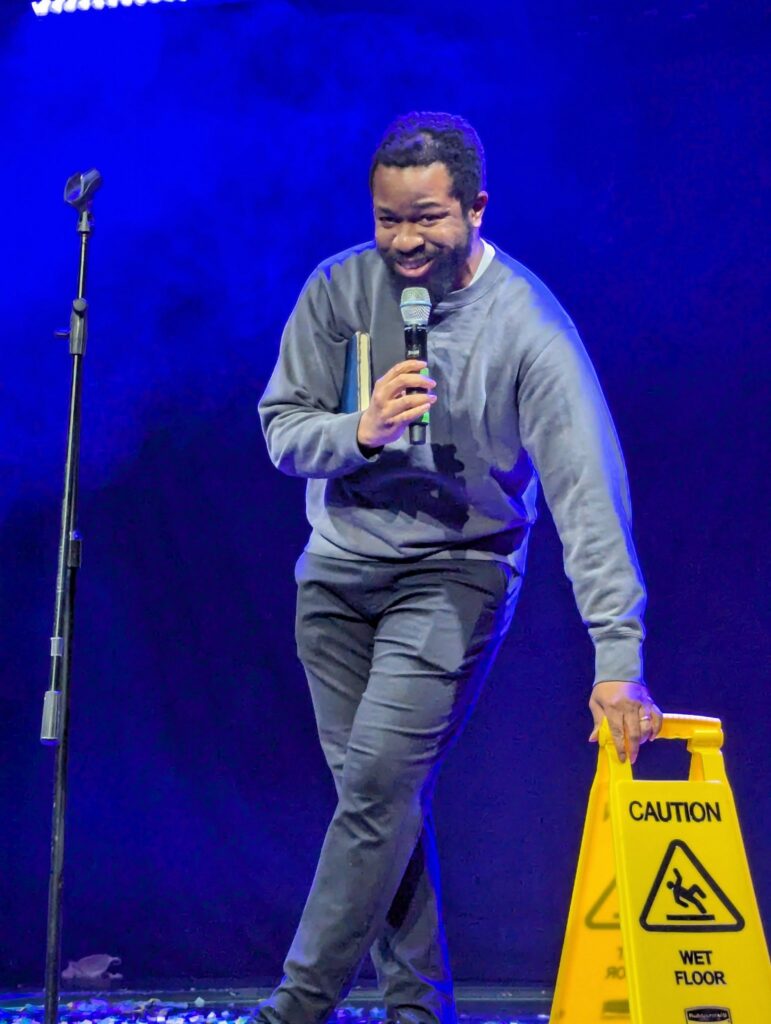 comedian leaning on a wet floor caution sign on stage with a sardonic expression