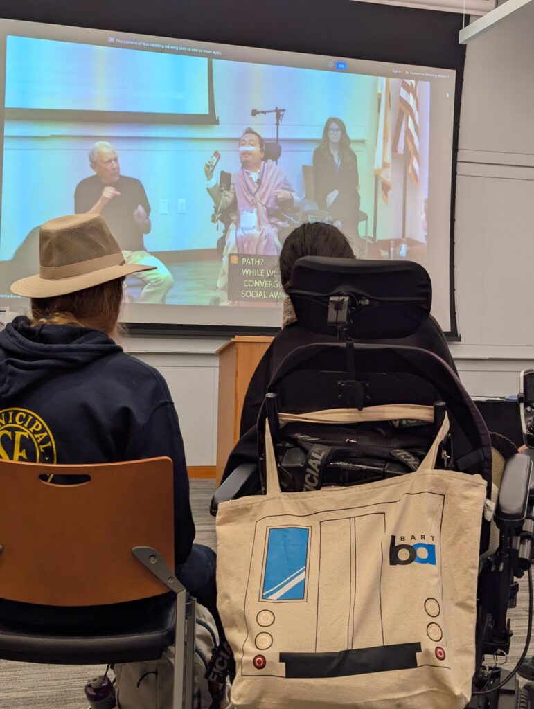 powerchair user with BART totebag on the back of her chair, and her seated partner, looking at conference speaker victor pineda
