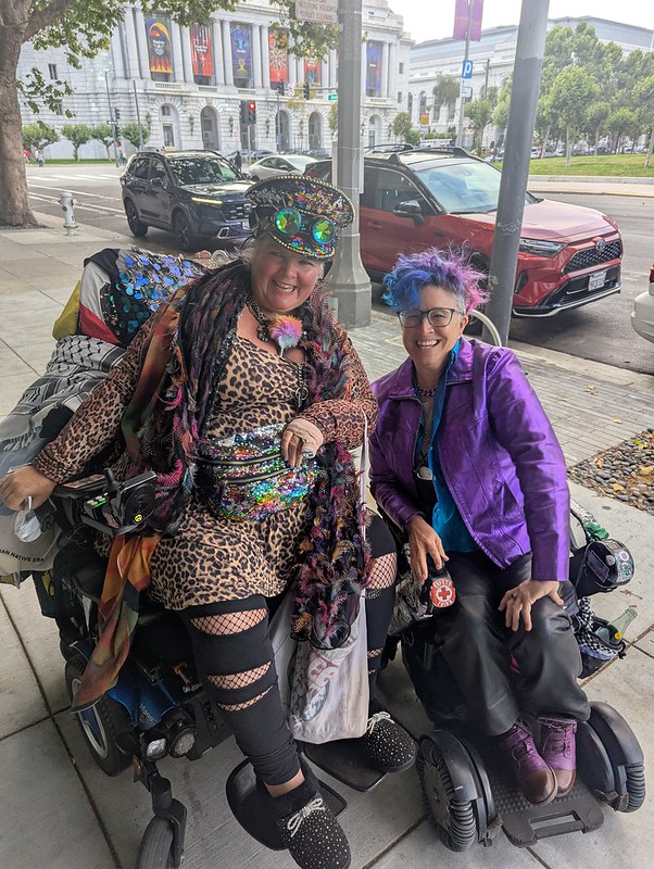 liz and maya in powerchairs outside the DCC, flamboyantly dressed